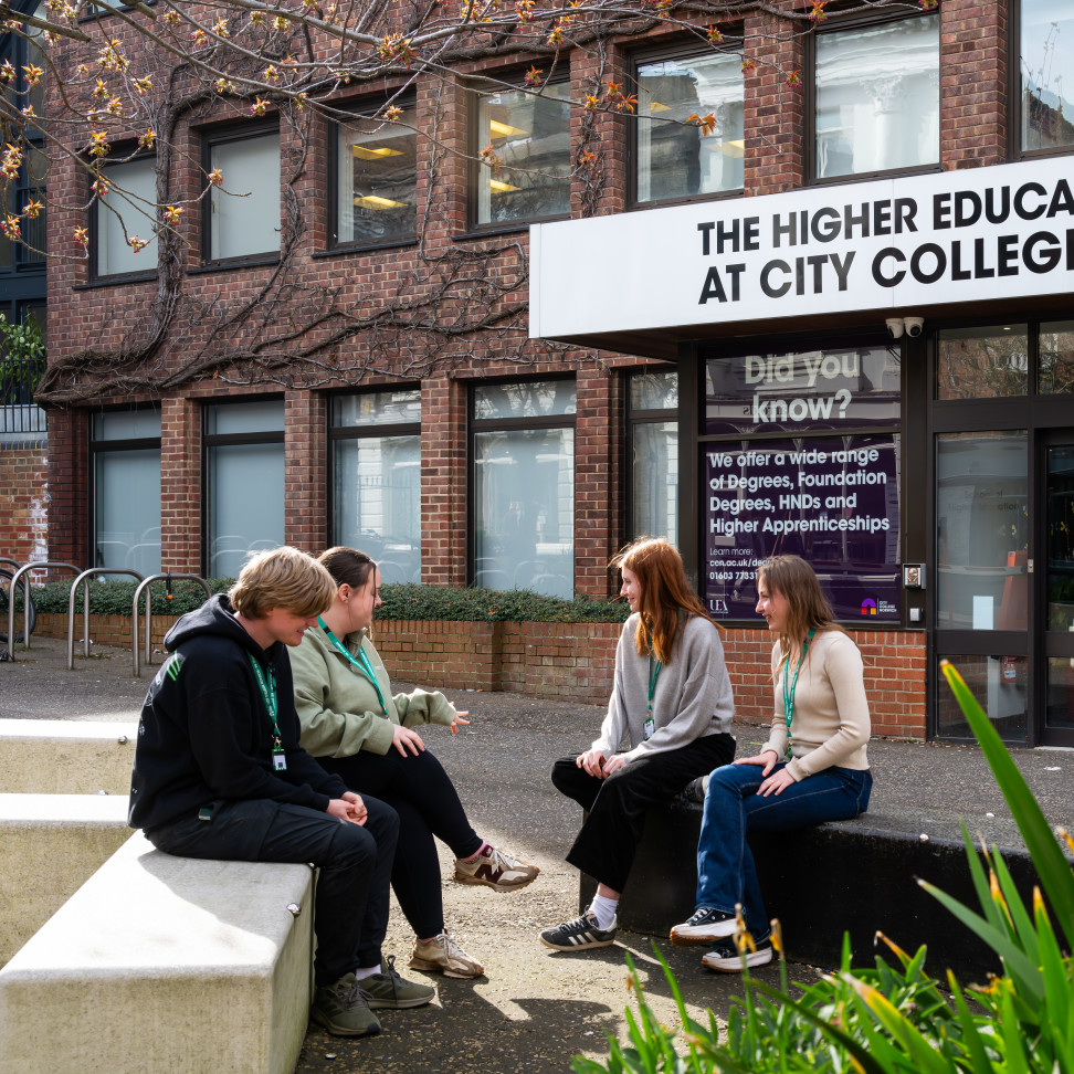 Four Higher Education students are sitting in a group outside Norfolk House, on benches.