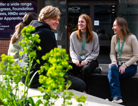 Four Higher Education students are chatting outside Norfolk House, on benches.