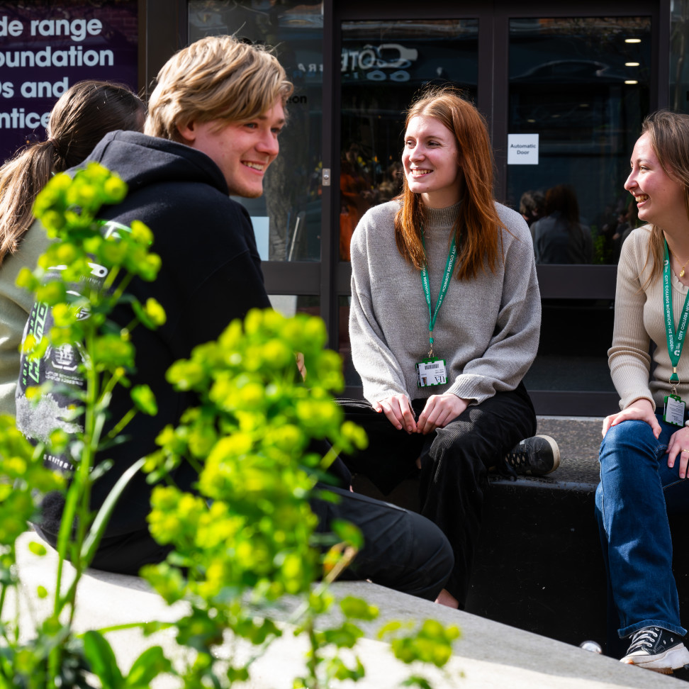 Four Higher Education students are chatting outside Norfolk House, on benches.